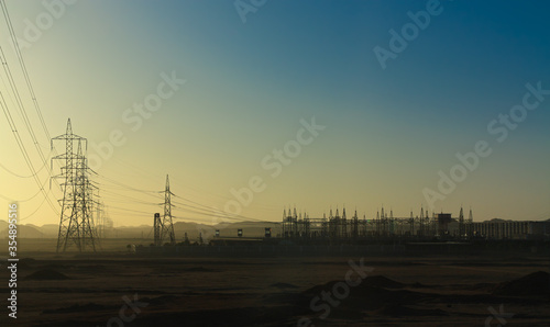 A power station far out in the desert in southern Egypt. The electricity pylons soar into the sky. In the background are the mountains in front of the desert.