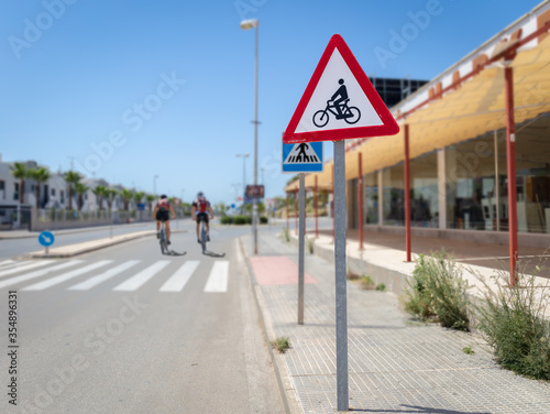 A triangular red and white sign warns of cyclists who could cross the road. In the background a crosswalk with a blue sign and two cyclists on the street.