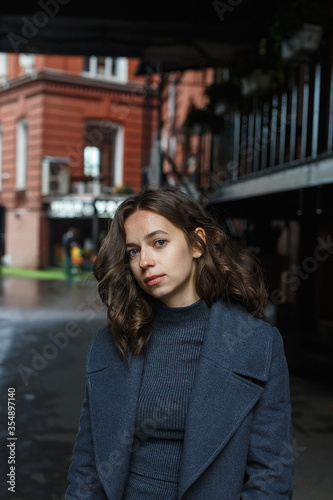 young thoughtful pretty girl in gray coat and turtleneck poses in front of red building. Street style portrait photo session of attractive female, urban photoshoot of beautiful elegant model