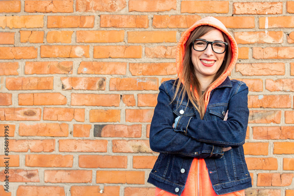 Smiling young woman in a denim jacket and glasses, arms crossed against a red brick wall