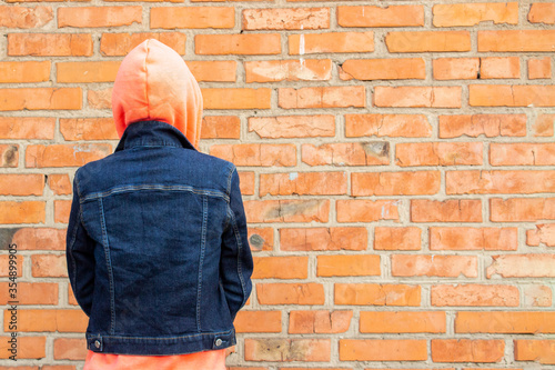 Young girl stands with her back against a red brick wall. Girl is dressed in a sweatshirt and denim jacket