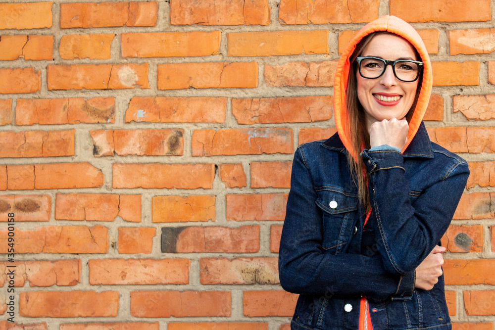 Happy young girl in glasses, dressed in a sweatshirt, wearing a hood on his head and in a denim jacket on a background of a red brick wall