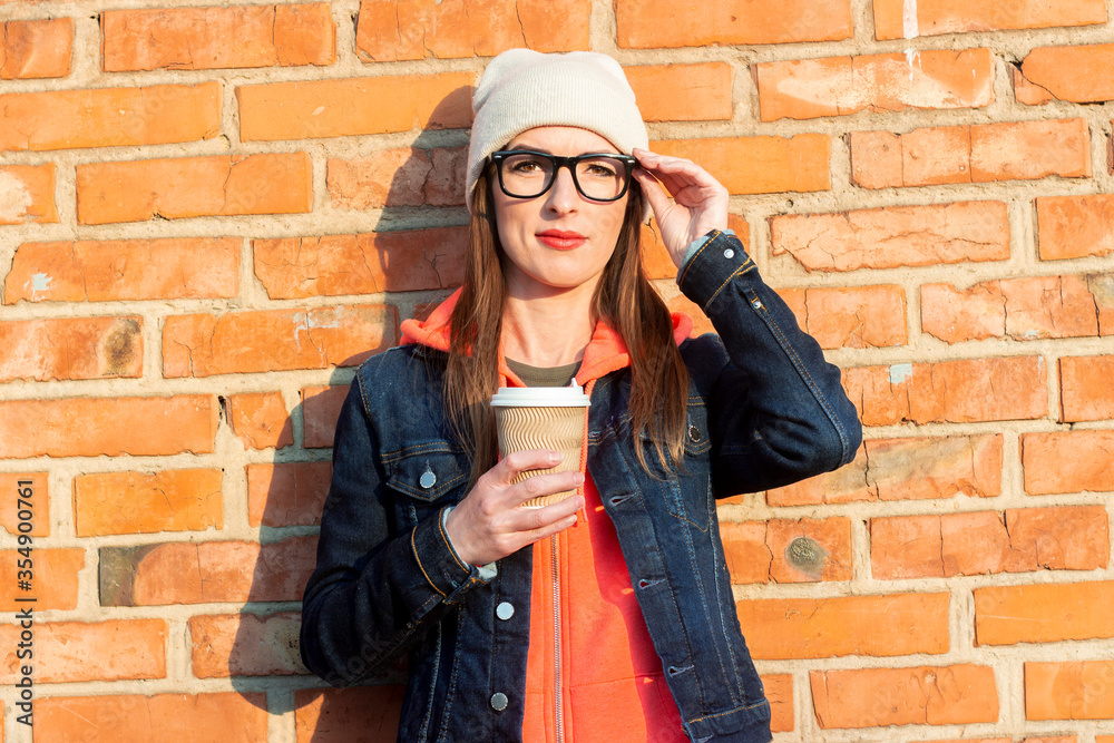 Young woman in glasses holds a paper cup with coffee on a brick wall background wearing a white hat and denim jacket Young woman in glasses holds a paper cup with coffee on a brick wall background wearing a white hat and denim jacket
