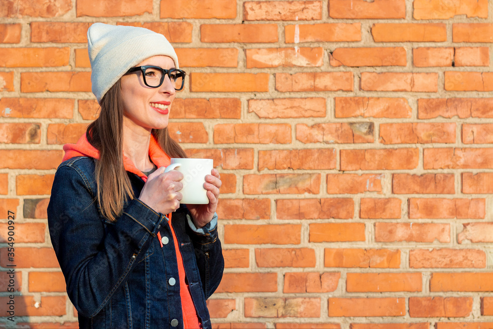Young smiling woman looking away in glasses holding a white cup on the background of brick wall wearing a white cap and a denim jacket.