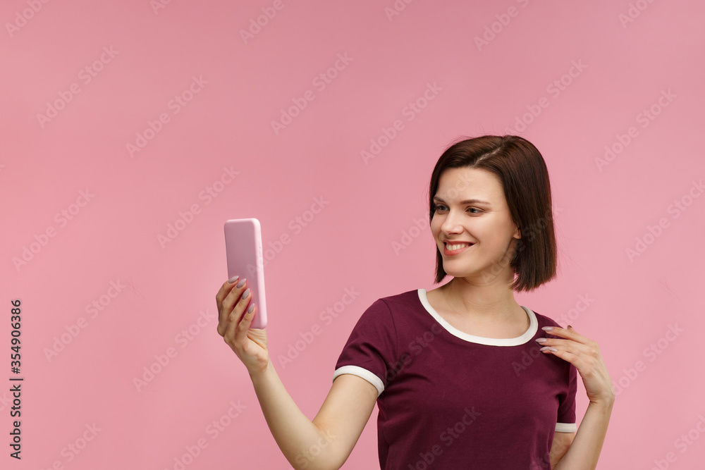 Funny young brunette woman making selfie. Smiling girl wearing t-shirt holding pink smartphone, making faces on camera, posing for selfie isolated on pink background. Funny young brunette woman making selfie. Smiling girl wearing t-shirt holding pink smartphone, making faces on camera, posing for selfie isolated on pink background.