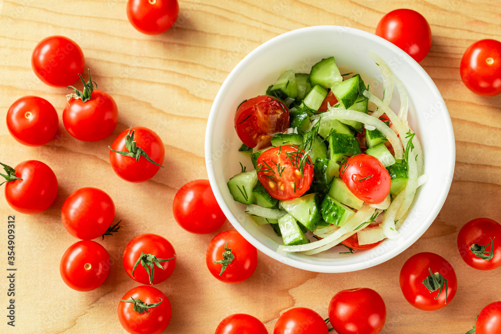 Cucumber, cherry tomatoes and onion salad in a deep white bowl on a light wooden Board. Next to the bowl is a lot of red cherry tomatoes Cucumber, cherry tomatoes and onion salad in a deep white bowl on a light wooden Board. Next to the bowl is a lot of red cherry tomatoes