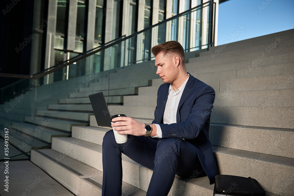 Fototapeta premium Young businessman sits on the stairs between high-rise buildings and is working with his laptop.
