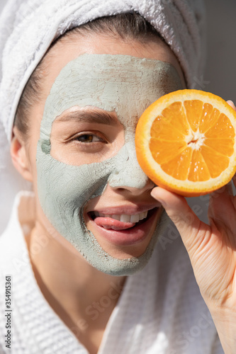 Young woman with facial clay mud mask on face holding orange slice covering eye and have fun