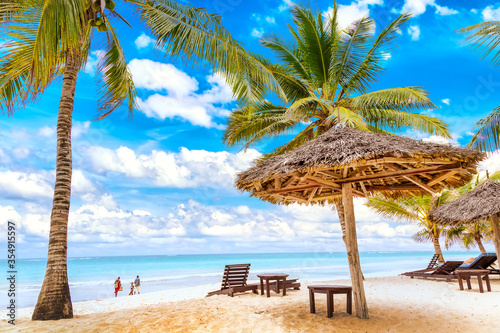 Sun loungers under umbrella and palms on the sandy beach by the ocean and cloudy sky. Vacation background. Idyllic beach landscape in Diani beach, Kenya, Africa