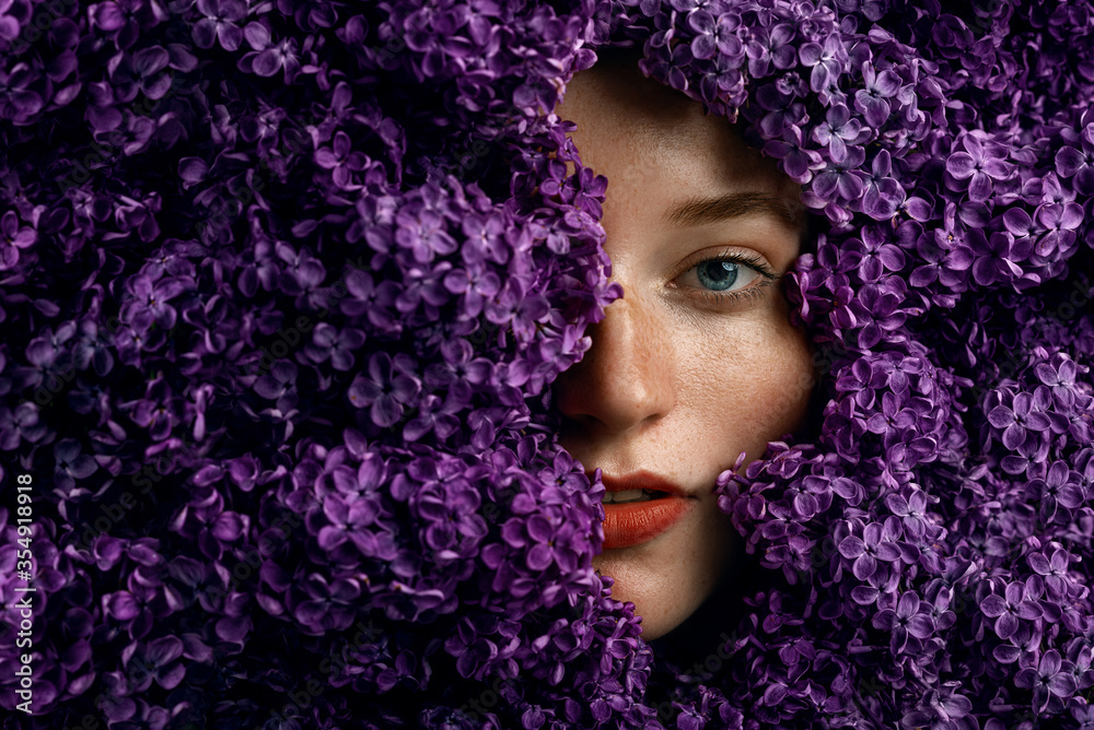 Woman hides her face in lilac flowers. Portrait of beautiful redhead girl with freckled skin, blue eyes. Copy, empty space for text Woman hides her face in lilac flowers. Portrait of beautiful redhead girl with freckled skin, blue eyes. Copy, empty space for text