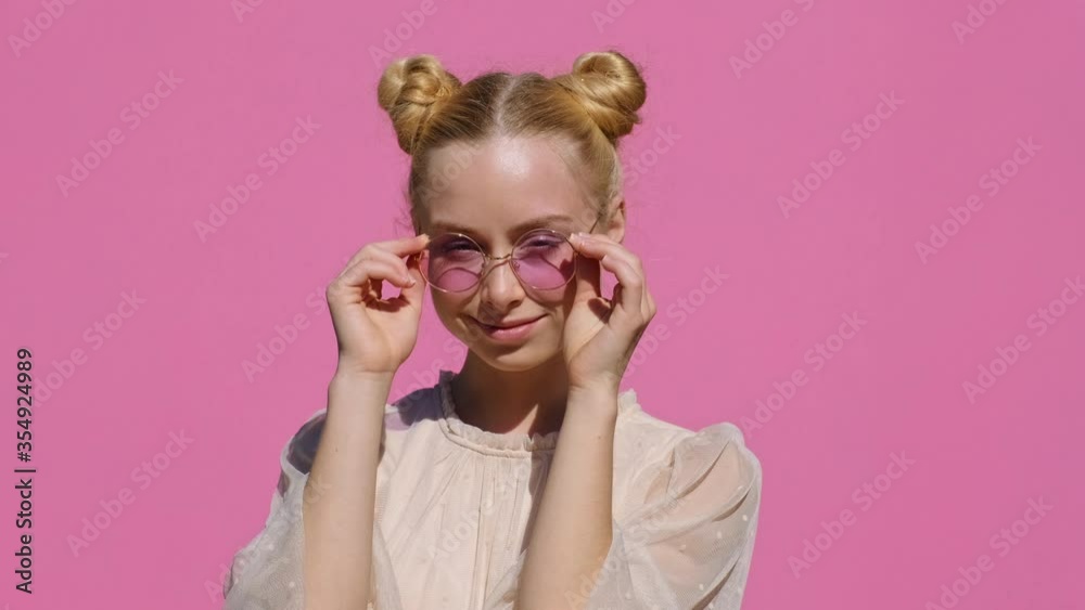Portrait of happy blonde teen girl in pink sunglasses touches hands with them smiling looking at camera on pink background slow motion in summer. Emotions of people. Positive mood. Portrait of happy blonde teen girl in pink sunglasses touches hands with them smiling looking at camera on pink background slow motion in summer. Emotions of people. Positive mood.