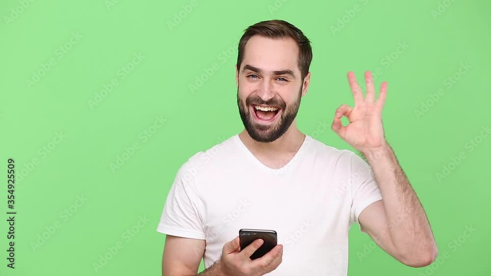 Confident happy young guy 20s in white t-shirt hold in hand using mobile cell phone send sms girlfriend showing ok okay gesture isolated on green chroma key background studio. People lifestyle concept Confident happy young guy 20s in white t-shirt hold in hand using mobile cell phone send sms girlfriend showing ok okay gesture isolated on green chroma key background studio. People lifestyle concept