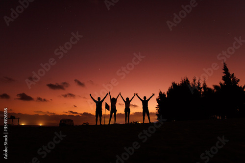 Group of four people in silhouette standing on beach with hands up and enjoying amazing sunset at tropical vacation. Happy friends spending time together during summer time.