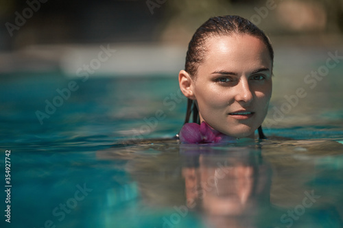 Stunning young girl with dark wet hair standing half water of swimming pool and looking directly on camera with blur background. Female tourist with enigmatic look during luxury relaxation