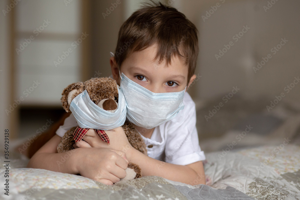 A boy in a mask laying on the bed and hugging a teddy bear in a mask. Image with selective focus
