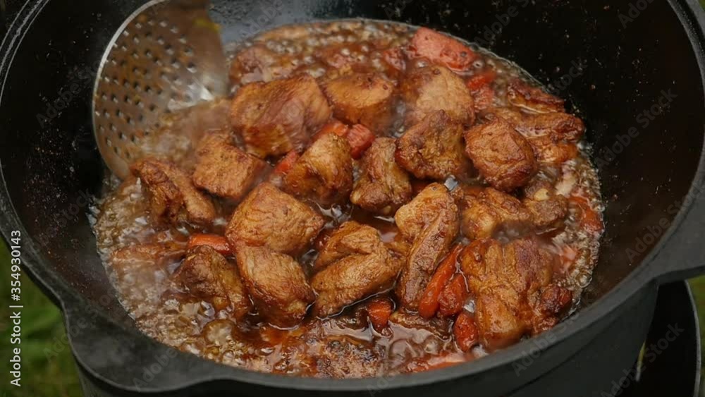 Cooking process, frying pork meat with carrots and onion in large cauldron on open fire. Close up, slow motion Cooking process, frying pork meat with carrots and onion in large cauldron on open fire. Close up, slow motion