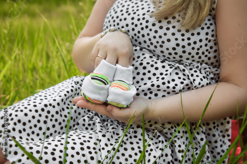 a pregnant girl in a polka-dot dress sits on the green grass in the forest and holds booties in the palm of her hand for her future son.
lmage with selective focus photo