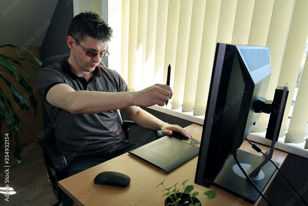 A man sits on a chair in front of a monitor and works on a project using a graphics tablet. He squints and focuses on the screen