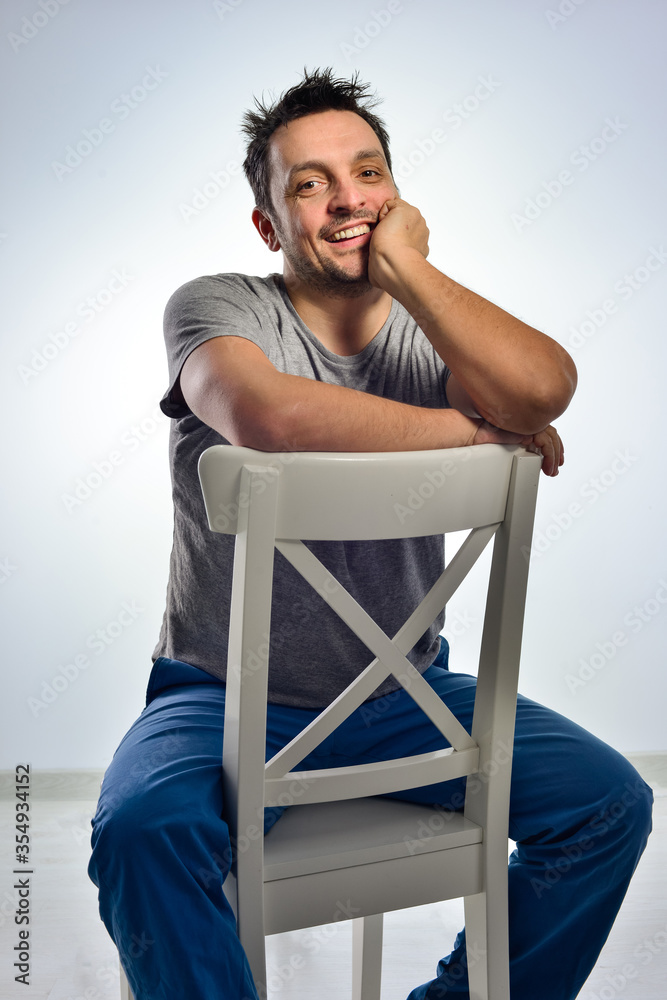 Young man dressed casual, sitting on a white chair, doing face expressions, isolated in white. Young man dressed casual, sitting on a white chair, doing face expressions, isolated in white.