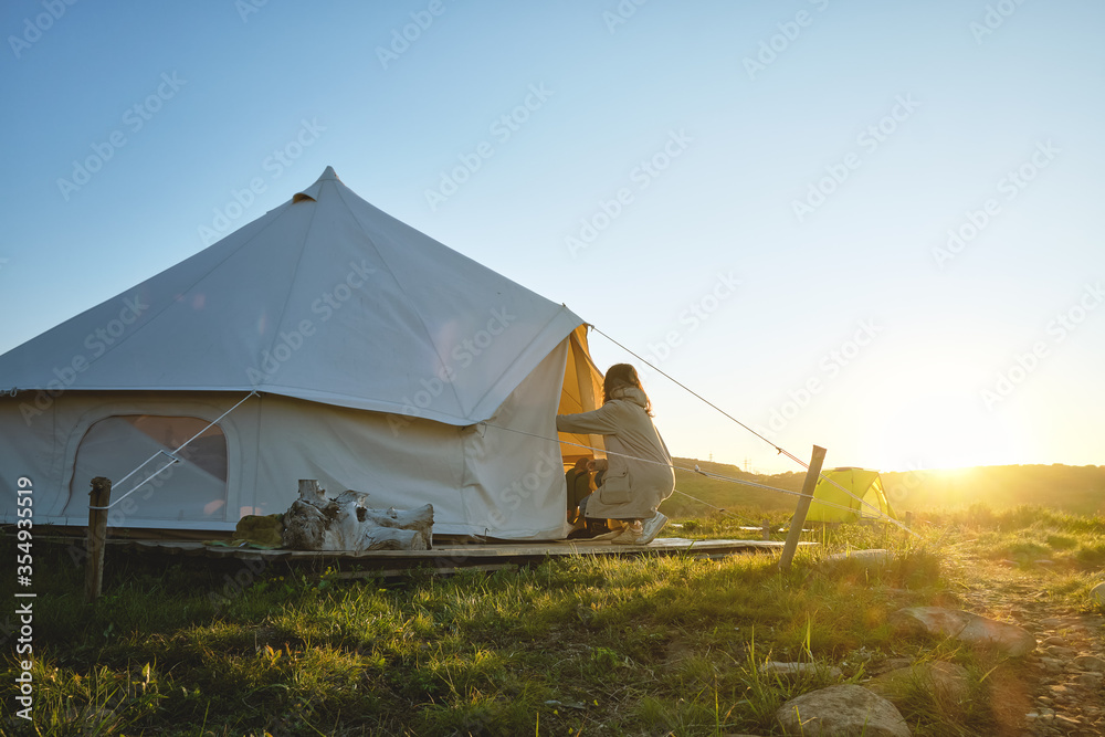 young girl opens a glamping tent in the middle of a green field in the sunset rays. Glamping and outdoor concept.