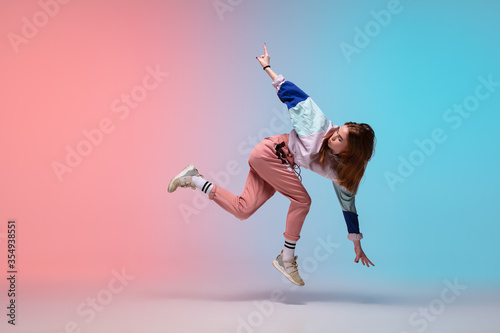 Fototapeta Naklejka Na Ścianę i Meble -  In jump. Beautiful girl dancing hip-hop in stylish clothes on colorful gradient background at dance hall in neon light. Youth culture, movement, style and fashion, action. Fashionable bright portrait.