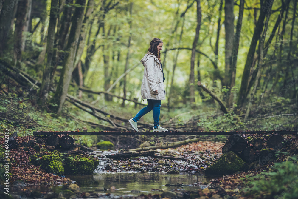 Side view of a young woman in a grey jacket and jeans walking over a wooden bridge over water in an enchanted mystical forest