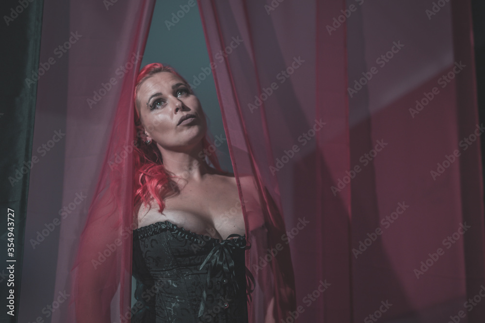 Woman in a sexy black corset or bustier going through transparent or see through curtains in front of a bed. Sexy redheaded curvy woman in lingerie spreading curtains and looking up