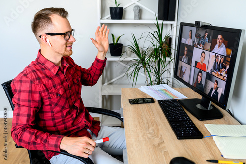 A young cheerful man greets with a gesture his coworkers in online video meeting, he sits at the office desk, a shelf with plants is on the background