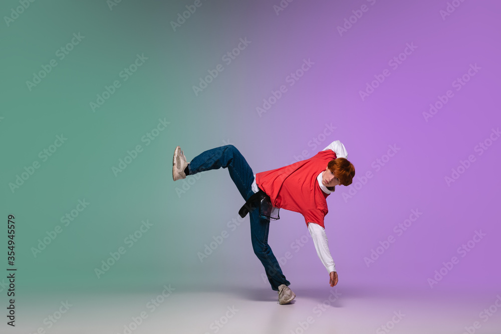 Beautiful redhead boy dancing hip-hop in stylish clothes on colorful gradient background at dance hall in neon light. Youth culture, movement, style and fashion, action. Fashionable bright portrait.