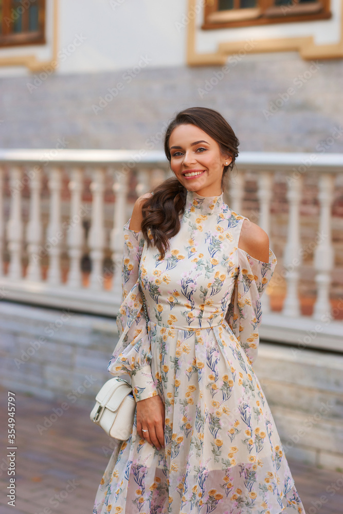 Pretty smiling young woman with long brunette hair wearing fashionable floral dress standing on street against balustrade on background. Happy female model in elegant stylish outfit posing outdoors. Pretty smiling young woman with long brunette hair wearing fashionable floral dress standing on street against balustrade on background. Happy female model in elegant stylish outfit posing outdoors.