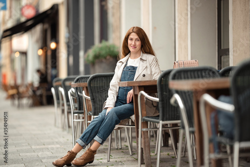 Beautiful young pregnant woman wearing sweater, jeans, grey coat sitting outside a cafe with a beautiful exterior in the summer.