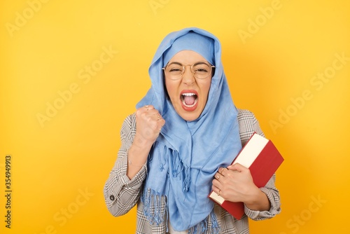 Caucasian young muslim woman wearing hijab, rejoicing her success and victory clenching her fists with joy.Lucky woman with hat being happy to achieve her aim and goals. Positive emotions, feelings. © Jihan