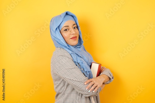 Image of cheerful pretty young muslim woman wearing hijab standing indoors with arms crossed. Looking and smiling at the camera. Confident girl. © Jihan