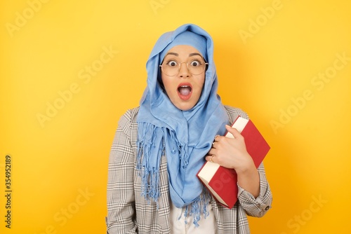 People and sudden reaction concept. Surprised young muslim woman wearing hijab, shrugs shoulders, looking sideways, being happy and excited about new plans with friends. Isolated over wall. © Jihan