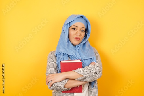 Waist up portrait of beautiful young muslim woman wearing hijab and casual clothes with positive expression, has crossed arms, feels happy and confident, isolated over bright background. © Jihan