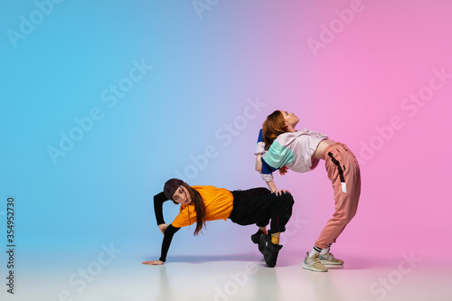 Flexible. Sportive girls dancing hip-hop in stylish clothes on colorful gradient background at dance hall in neon light. Youth culture, movement, style and fashion, action. Fashionable portrait.