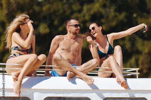 Smiling young people in sunglasses taking photos on mobile phone and  relaxing on top of hippie bus. Cheerful friends in swimsuits taking fun during sunny days on vacation.