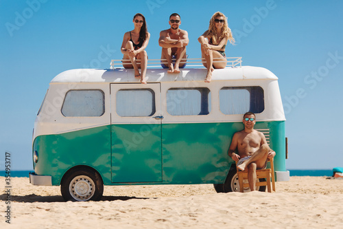 Canvas Print Group of four smiling travelers in sunglasses and swimsuits taking fun near retro hippie car on beach