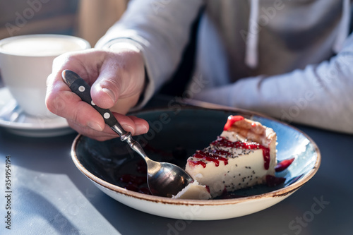 Delicious cheesecake breaks off with a spoon from a large piece on a beautiful plate, on a blurred background a cup of cappuccino.