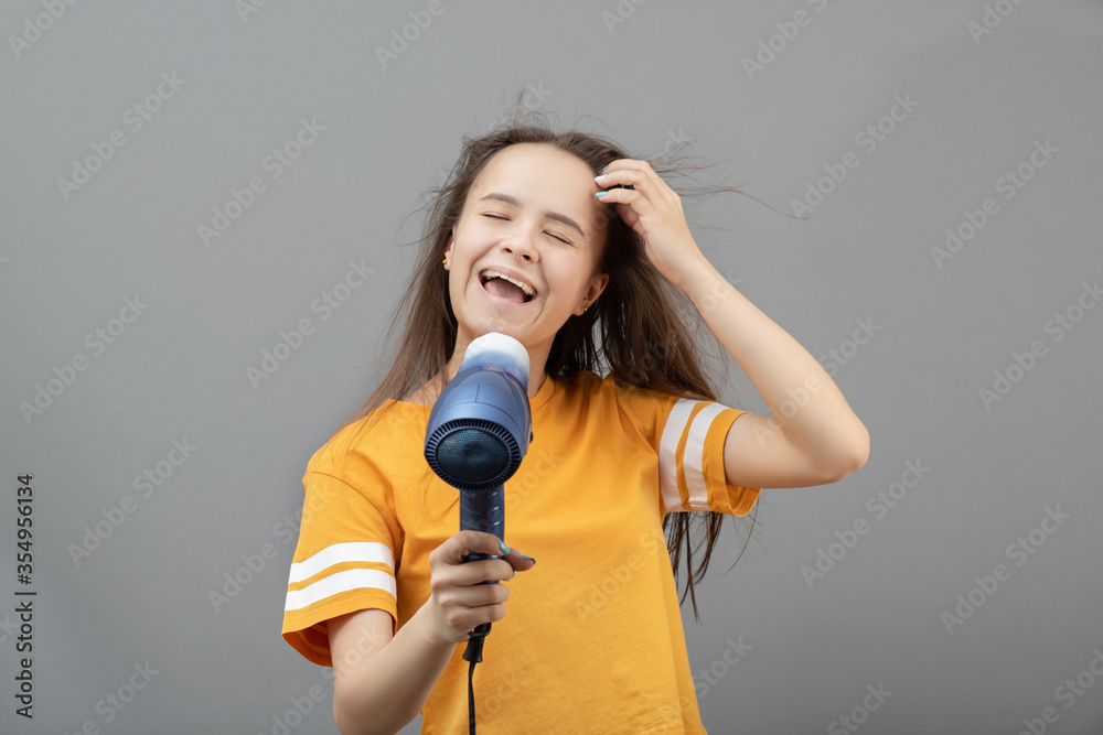 Young caucasian woman dries hair with a hairdryer on a gray background, has fun, sings like a microphone Young caucasian woman dries hair with a hairdryer on a gray background, has fun, sings like a microphone