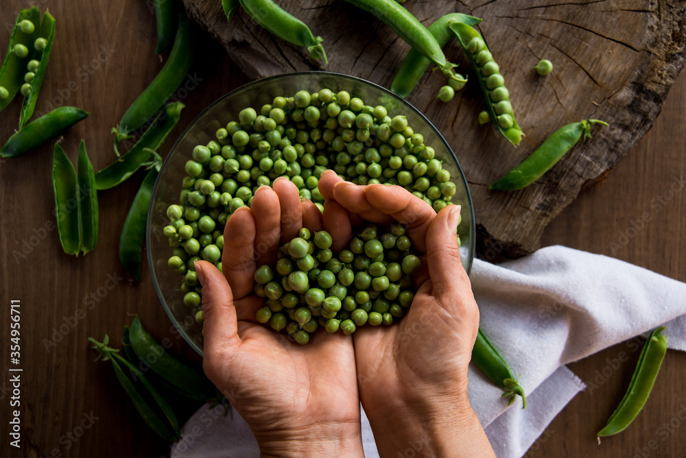 White female hands holding a handful green peas on a wooden table. In the table there are a crystal bowl full of green peas and a linen cloth. Rustic style. Top view.
