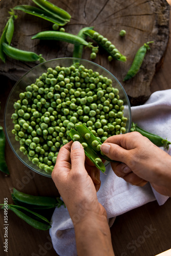 White female hands cleaning green peas on a wooden table. In the table there are a crystal bowl full of green peas and a linen cloth. Rustic style. Top view.