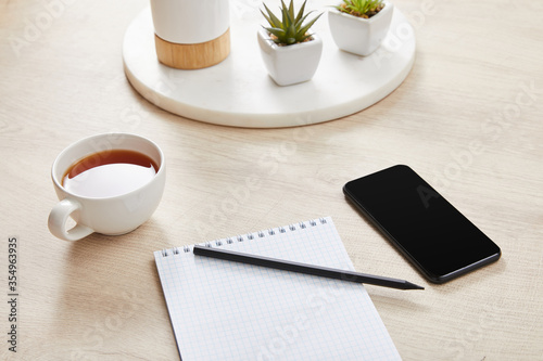 green plants, cup of tea and blank notebook with pencil near smartphone on wooden surface