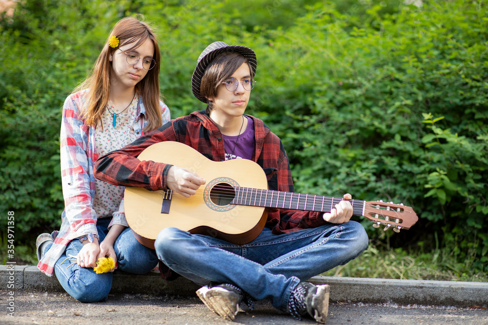 a pair of hipie boy and teen girl play guitar sitting on the sidewalk Asking For Money Donation