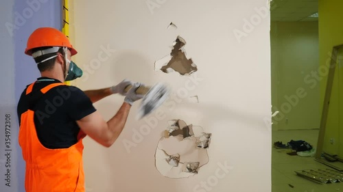 Worker in uniform and respirator destroys inner wall sledge hammer, back view. Demolition work and rearrangement concept. Wall made of gypsum cardboard being destroyed. 4 k video