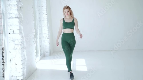 young sexy fitness girl in leggings and a T-shirt with a beautiful athletic body goes to the camera with her hands up. Blonde woman posing at camera. Slow motion. White studio background