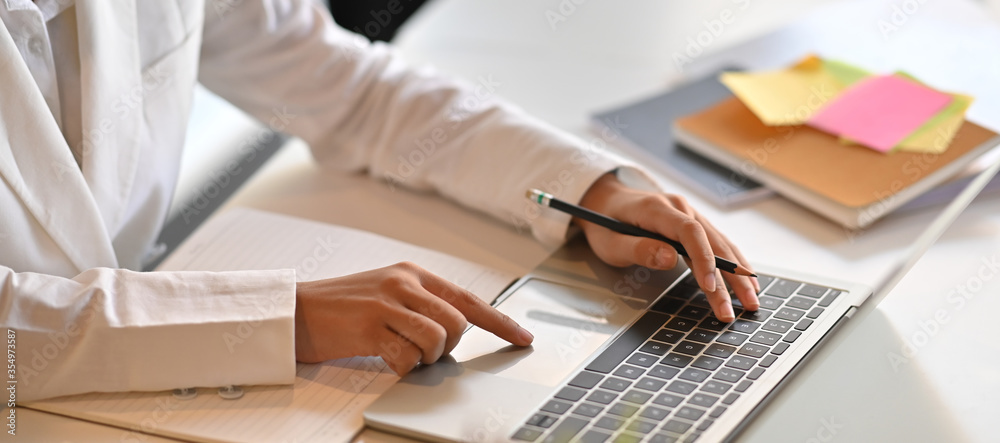 Hands of a businesswoman are taking notes while working with a computer laptop that putting on a white working desk surrounded by office equipment.
