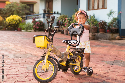 Playful Pretty Indian girl child infant toddler wearing a hair band  playing with a cycle tricycle. Kid giving joyful expressions