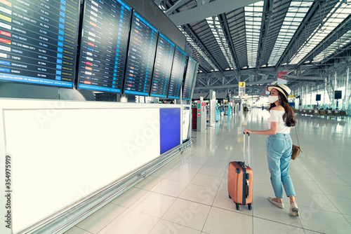 Asian female wearing face mask with suitcase checking flight cancellation status on airport information board in empty airport. airline bankrupt, airline crisis, coronavirus or new normal concept photo