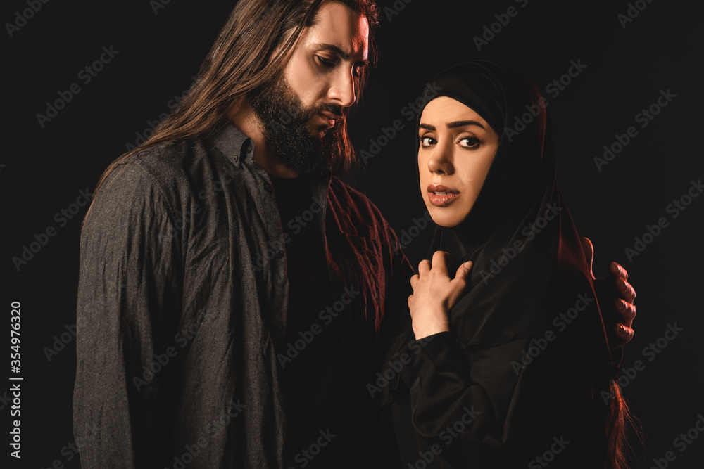 Muslim woman in hijab looking at camera near husband isolated on black Muslim woman in hijab looking at camera near husband isolated on black